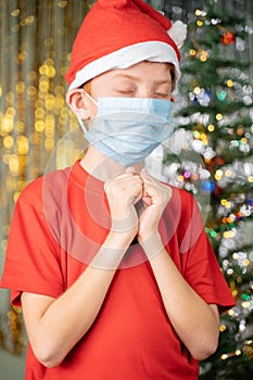 Kid in medical mask and Santa hat praying for santa on Christmas decorated background for gifts.