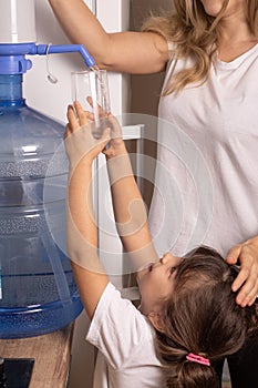 Kid and mother drinks clean water from a transparent glass from manual drinking water pump at home.