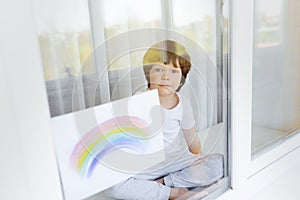 Kid painting rainbow during Covid-19 quarantine at home. Boy near window. Stay at home Social media campaign for coronavirus