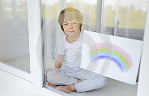 Kid painting rainbow during Covid-19 quarantine at home. Boy near window. Stay at home Social media campaign for coronavirus