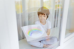 Kid painting rainbow during Covid-19 quarantine at home. Boy near window. Stay at home Social media campaign for coronavirus