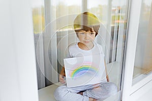 Kid painting rainbow during Covid-19 quarantine at home. Boy near window. Stay at home Social media campaign for coronavirus