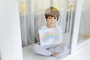 Kid painting rainbow during Covid-19 quarantine at home. Boy near window. Stay at home Social media campaign for coronavirus