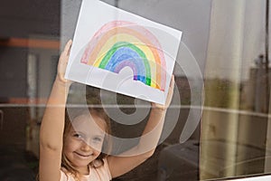 Kid painting rainbow during Covid-19 quarantine at home. Girl near window. Stay at home Social media campaign for coronavirus