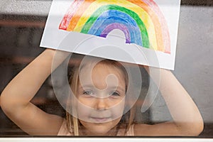 Kid painting rainbow during Covid-19 quarantine at home. Girl near window. Stay at home Social media campaign for coronavirus