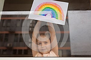 Kid painting rainbow during Covid-19 quarantine at home. Girl near window. Stay at home Social media campaign for coronavirus