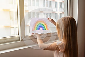 Kid painting rainbow during Covid-19 quarantine at home. Girl near window. Stay at home Social media campaign for coronavirus