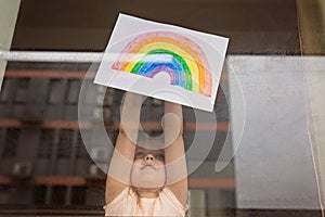 Kid painting rainbow during Covid-19 quarantine at home. Girl near window. Stay at home Social media campaign for coronavirus