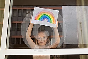 Kid painting rainbow during Covid-19 quarantine at home. Girl near window. Stay at home Social media campaign for coronavirus