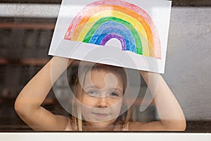 Kid painting rainbow during Covid-19 quarantine at home. Girl near window. Stay at home Social media campaign for coronavirus