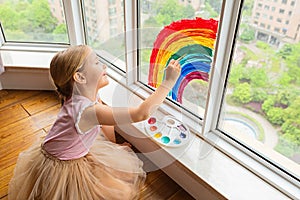 Kid painting rainbow during Covid-19 quarantine at home. Girl near window. Stay at home Social media campaign for coronavirus