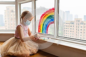 Kid painting rainbow during Covid-19 quarantine at home. Girl near window. Stay at home Social media campaign for coronavirus