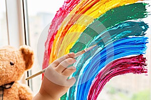 Kid painting rainbow during Covid-19 quarantine at home. Girl near window. Stay at home Social media campaign for coronavirus