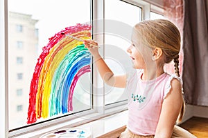 Kid painting rainbow during Covid-19 quarantine at home. Girl near window. Stay at home Social media campaign for coronavirus