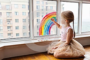 Kid painting rainbow during Covid-19 quarantine at home. Girl near window. Stay at home Social media campaign for coronavirus