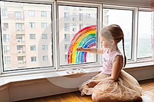 Kid painting rainbow during Covid-19 quarantine at home. Girl near window. Stay at home Social media campaign for coronavirus