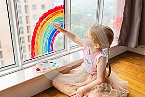 Kid painting rainbow during Covid-19 quarantine at home. Girl near window. Stay at home Social media campaign for coronavirus