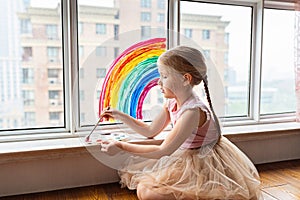 Kid painting rainbow during Covid-19 quarantine at home. Girl near window. Stay at home Social media campaign for coronavirus