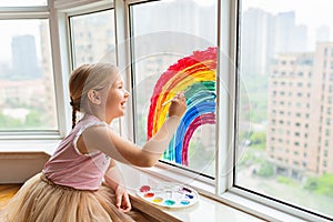 Kid painting rainbow during Covid-19 quarantine at home. Girl near window. Stay at home Social media campaign for coronavirus