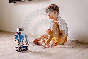 Kid programming robot from plastics details and programmed on tablet, robotics. Curly boy lies on floor with tablet