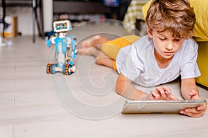 Kid programming robot from plastics details and programmed on tablet, robotics. Curly boy lies on floor with tablet