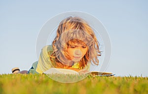 Kid read book in park. Child boy reading book laying on grass on grass and sky background with copy space outdoor.