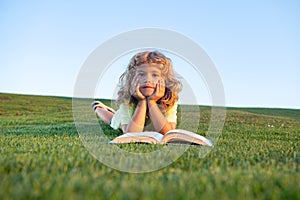 Kid reading book laying on grass on grass and sky background with copy space. Funny child boy reading the book in spring