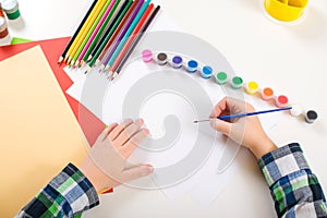 Kid`s drawing. Art background with colorful pencils, paints and empty paper. Kid hands with brush on a paper. Children`s