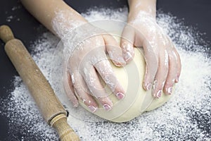 Kid`s hands, some flour, wheat dough and rolling pin on the black table. Children hands making the rye dough for backing bread.