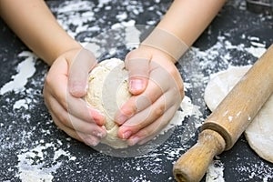 Kid`s hands, some flour, wheat dough and rolling pin on the black table. Children hands making the rye dough for backing bread. S