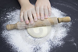 Kid`s hands, some flour, wheat dough and rolling pin on the black table. Children hands making the rye dough for backing bread.