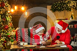 Kid Santa Claus enjoying in served gingerbread cake and milk. Santa Father and son having fun near Christmas tree and