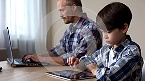 Kid scrolling on tablet, father working on laptop computer, lack of attention