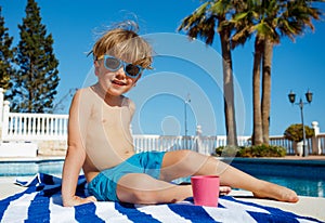 Kid in shades grins on towel by pool at sunny weather.
