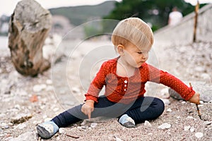 Kid sits on a pebble beach and digs a hole in the ground with a stick