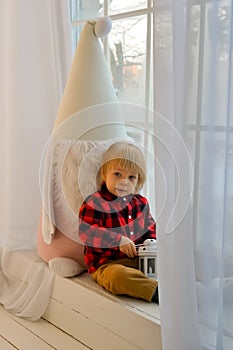 The kid sits on the windowsill with a Christmas gnome and holds a Christmas lantern and a New Year& x27;s gift, box in