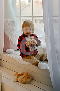 The kid sits on the windowsill with a Christmas gnome and holds a Christmas lantern and a New Year& x27;s gift, box in