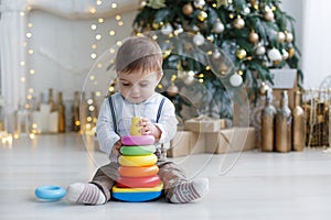 The kid, sitting on the floor near a smart Christmas tree, collects a multicolored pyramid.