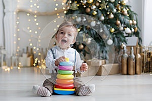 The kid, sitting on the floor near a smart Christmas tree, collects a multicolored pyramid.