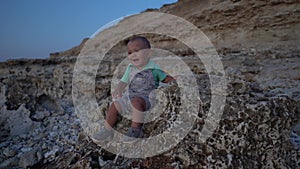 Kid sitting on the stones on the sea coast at sunset . Boy talking and fidgeting