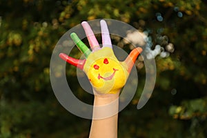 Kid with smiling face drawn on palm in green park. School holidays