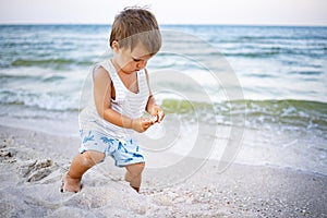 A kid in a striped T-shirt collects pebbles and shells on the coast on a summer evening