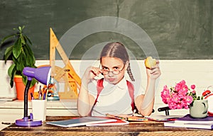 Kid student in school. Schoolgirl sit desk chalkboard background. Girl small child eating apple snack. School break