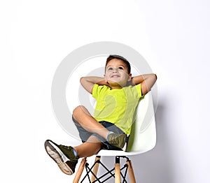 Kid in sunglasses, yellow t-shirt, denim shorts, khaki sneakers. Smiling showing victory sign, sitting on chair.  on white