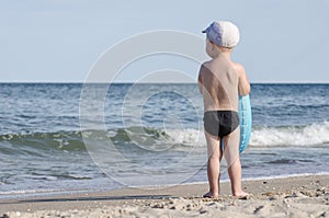 Kid in swimming trunks with a swimming circle stands on the shore of the sea back