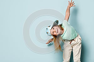 Kid in t-shirt, pants and cap printed by pineapples. She leaned forward with her arms wide apart, smiling, posing on turquoise