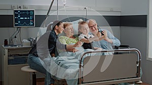 Kid taking selfie on smartphone with ill patient and family at visit
