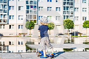 Kid toddler riding his scooter in a city. Summertime fun activity. Caucasian white boy is wearing a hat.