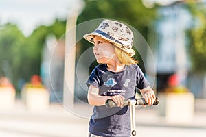 Kid toddler riding his scooter in a city. Summertime fun activity. Caucasian white boy is wearing a hat.