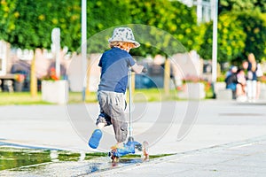 Kid toddler riding his scooter in a city. Summertime fun activity. Caucasian white boy is wearing a hat. Back view